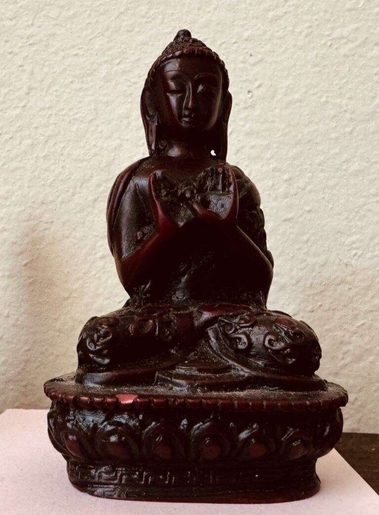 Photo of a small statue of Buddha. His eyes are closed, he sits in the double lotus position, both hands held at chest height, palms outward.